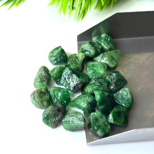 May include: A collection of raw, uncut emerald gemstones. The rough emeralds are a deep, vibrant green colour, with a natural, unpolished appearance. The gemstones are scattered on a white surface.