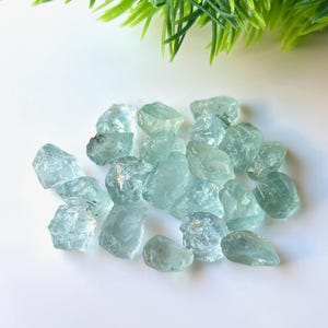 May include: A collection of light blue aquamarine rough stones. The stones are various sizes and shapes, with some having a more translucent appearance than others.