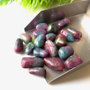 Peut inclure: Un assortiment de pierres précieuses polies dans un plateau métallique. Les pierres présentent un mélange de couleurs, notamment du rouge profond, du sarcelle et des touches de vert. Les pierres ont un aspect lisse et poli. Le fond est blanc avec une touche de vert.