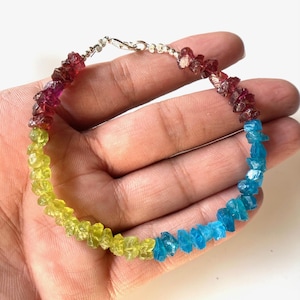 May include: A multi-colored gemstone bracelet with red, yellow, and blue stones. The bracelet is made of small, irregular shaped stones and is strung on a silver chain.