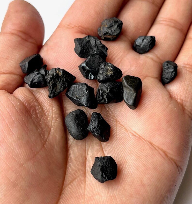 May include: A collection of small, black, rough, and irregularly shaped stones. The stones are scattered on a person's hand.