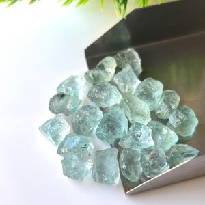 May include: A pile of light blue aquamarine rough gemstones in a metal scoop. The gemstones are various sizes and shapes.