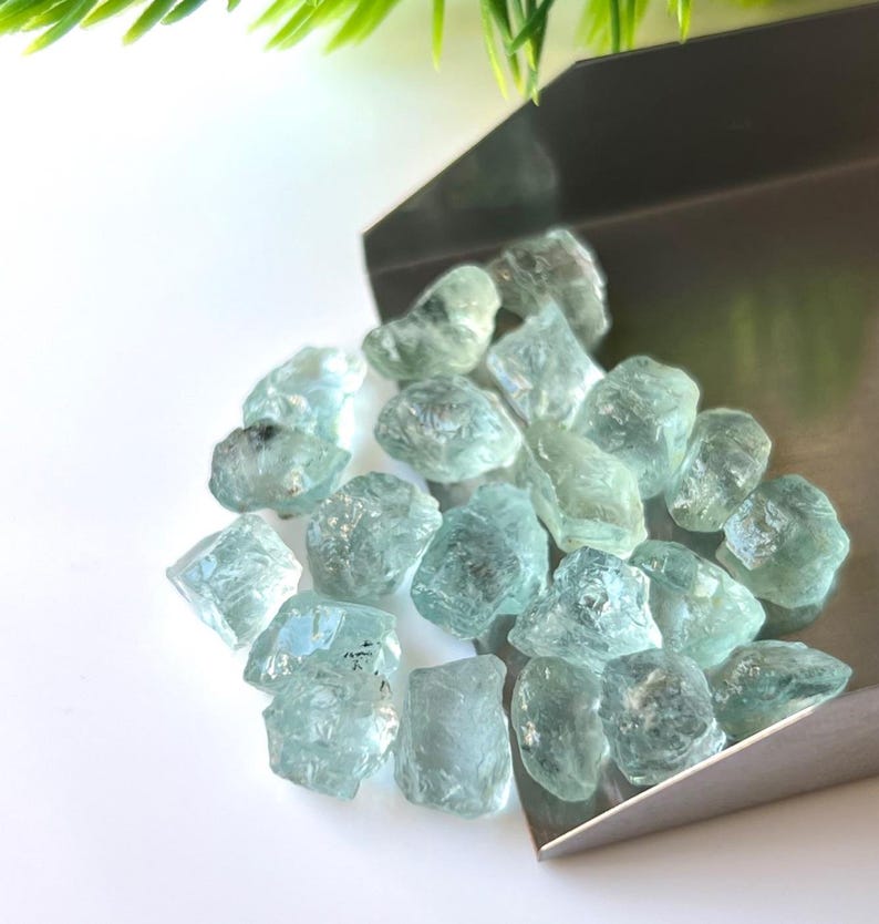 May include: A pile of light blue aquamarine rough gemstones in a metal scoop. The gemstones are various sizes and shapes.