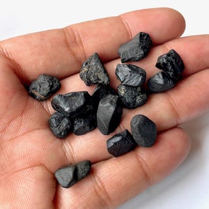 May include: A collection of black, rough, and irregularly shaped gemstones. The stones are various sizes and have a matte finish.