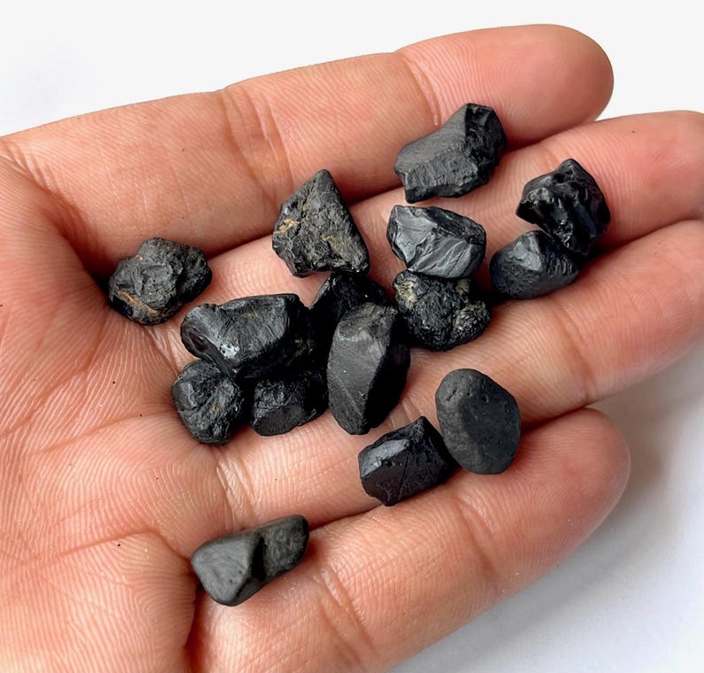 May include: A collection of black, rough, and irregularly shaped gemstones. The stones are various sizes and have a matte finish.