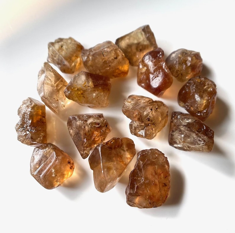 Puede incluir: Un conjunto de piedras preciosas de citrino en bruto y sin cortar. Las piedras, de tama&ntilde;os y formas variadas, presentan un color marr&oacute;n &aacute;mbar c&aacute;lido y una calidad transl&uacute;cida. Las piedras est&aacute;n dispuestas sobre una superficie blanca.
