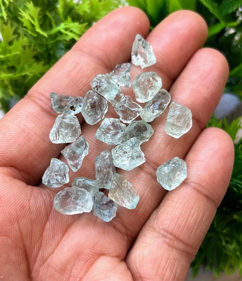 May include: A collection of small, light blue aquamarine crystals in a hand. The crystals are rough and have a natural, unpolished look.