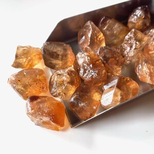 Puede incluir: Colecci&oacute;n de gemas de citrino en bruto, sin cortar. Las piedras, de color &aacute;mbar c&aacute;lido, var&iacute;an en tama&ntilde;o y forma. Se muestran en una peque&ntilde;a cuchara plateada sobre un fondo blanco. Los cristales de citrino tienen una textura natural y rugosa.