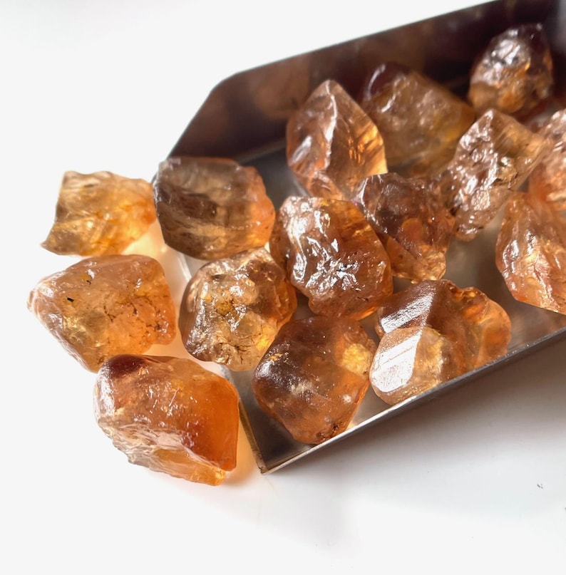 Puede incluir: Colecci&oacute;n de gemas de citrino en bruto, sin cortar. Las piedras, de color &aacute;mbar c&aacute;lido, var&iacute;an en tama&ntilde;o y forma. Se muestran en una peque&ntilde;a cuchara plateada sobre un fondo blanco. Los cristales de citrino tienen una textura natural y rugosa.