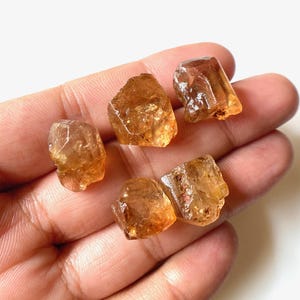 Puede incluir: Cinco cristales de piedras preciosas en bruto, translúcidos, facetados, de color naranja-marrón.