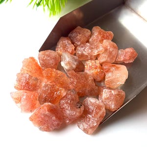 May include: A collection of raw, unpolished sunstone crystals with a distinct orange and reddish-brown hue, featuring natural sparkling inclusions. These rough, irregular mineral stones are gathered together inside a metallic silver-toned scoop.