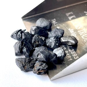 May include: A close-up of a pile of black obsidian stones, some with a rough, jagged texture, others with a smoother surface. The stones are spilling out of a silver metal scoop with the text "K.K. Gems Trading Jaipur Tel: +91-141-38893 05 6181" engraved on the side.