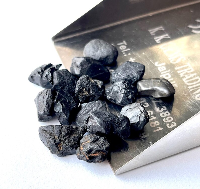 May include: A close-up of a pile of black obsidian stones, some with a rough, jagged texture, others with a smoother surface. The stones are spilling out of a silver metal scoop with the text "K.K. Gems Trading Jaipur Tel: +91-141-38893 05 6181" engraved on the side.