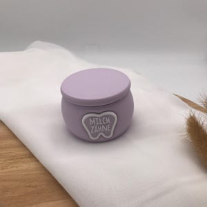 Könnte beinhalten: Ein kleiner, lilafarbener Behälter mit Deckel, auf einem weißen Tuch. Der Behälter hat ein weißes Emblem mit den Worten "MILCH ZAHNE" in einem zahnförmigen Design. Wahrscheinlich zur Aufbewahrung von Milchzähnen.