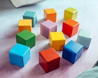 May include: A collection of twelve brightly colored wooden blocks arranged in a circle. The blocks are painted in various colors, including blue, green, yellow, orange, red, and purple. The blocks are approximately 1.5 inches square.