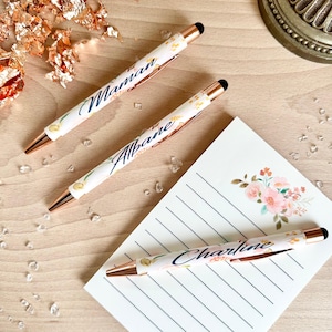 Op de afbeelding: Drie gepersonaliseerde pennen met bloemenmotieven en gouden accenten. De pennen hebben de namen "Maman", "Albane" en "Charline" erop geschreven.