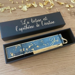 Op de afbeelding: Een zwarte doos met een gouden inscriptie die "La lecture est l'apothéose de l'écriture" luidt en een blauw en gouden bladwijzer met een gouden kwast en de naam "Zoé" erop.