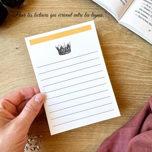 May include: A white lined notepad with a gold header and a crown illustration. The notepad has the text "Pour les lecteurs qui écrivent entre les lignes" at the top. The notepad is held by a hand and is on a wooden surface.