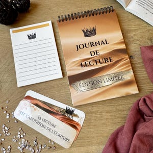 Peut inclure: Ensemble de papeterie comprenant un journal à spirales avec le texte « JOURNAL DE LECTURE » et « ÉDITION LIMITÉE », un bloc-notes et un marque-page. Le journal et le bloc-notes présentent un motif de couronne. Les articles sont disposés sur une surface en bois.
