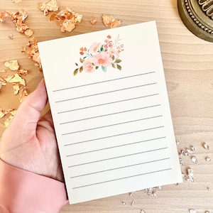 May include: A cream-coloured lined stationery card with a floral design in the upper corner. The watercolour-style flowers are pink, peach, and green. The card is held by a person.