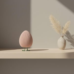 Puede incluir: Un objeto decorativo rosa en forma de huevo con estrías verticales, sobre patas verdes. Junto a él, un pequeño jarrón blanco contiene hierba ornamental seca. Los objetos están sobre una estantería de color claro.