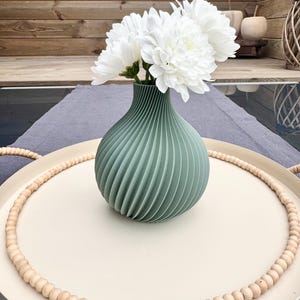 Op de afbeelding: Een saliegroene vaas met een geribbeld ontwerp houdt een boeket witte bloemen vast. De vaas staat op een crèmekleurig dienblad met een houten kralenrand. De lade staat op een grijs oppervlak, met een houten achtergrond.