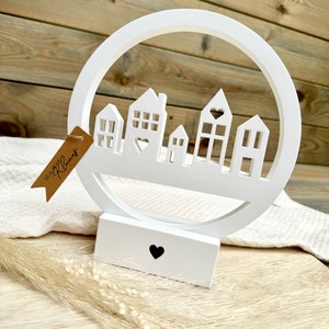 Könnte beinhalten: Weiße Holz-Dekoration für zu Hause mit einem runden Ausschnitt und einer Silhouette von Häusern mit Herzen. Die Basis der Dekoration hat einen kleinen Herz-Ausschnitt.