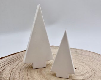 May include: Two white ceramic Christmas tree ornaments on a wooden slice. The trees are simple geometric shapes with a flat base.