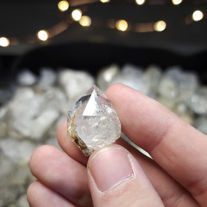 Peut inclure: Un cristal de quartz clair, facett&eacute;, &agrave; double terminaison avec une pointe &agrave; chaque extr&eacute;mit&eacute;. Le cristal est tenu dans une main, et l'arri&egrave;re-plan est flou.