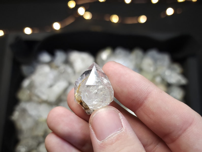 Peut inclure: Un cristal de quartz clair, facett&eacute;, &agrave; double terminaison avec une pointe &agrave; chaque extr&eacute;mit&eacute;. Le cristal est tenu dans une main, et l'arri&egrave;re-plan est flou.