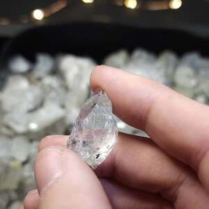 Peut inclure: Un cristal clair et facett&eacute; avec une pointe pointue. Le cristal est tenu dans une main, et il y a d'autres cristaux en arri&egrave;re-plan.