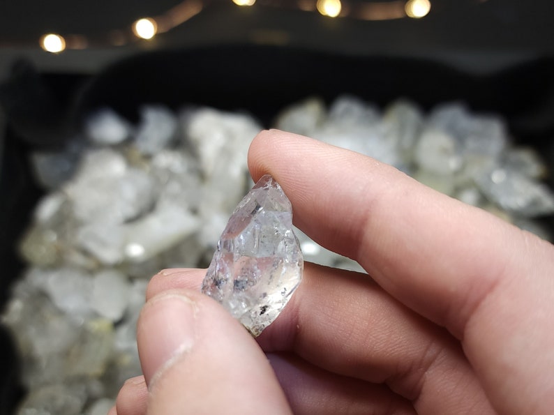 Peut inclure: Un cristal clair et facett&eacute; avec une pointe pointue. Le cristal est tenu dans une main, et il y a d'autres cristaux en arri&egrave;re-plan.