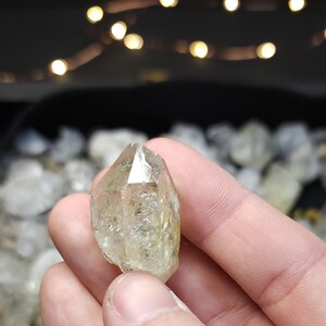 Peut inclure: Un cristal de quartz clair et facett&eacute; avec une teinte jaune. Le cristal est tenu dans une main, avec d'autres cristaux visibles en arri&egrave;re-plan.