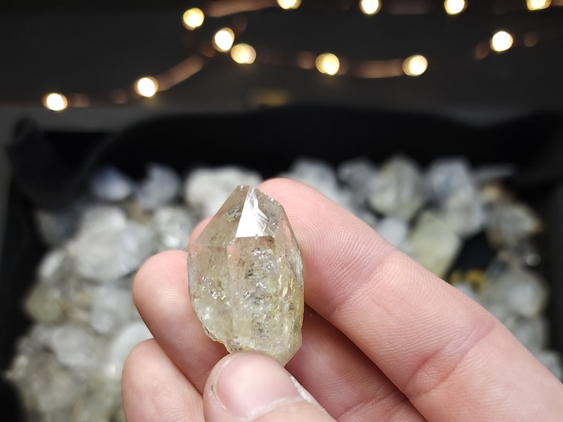 Peut inclure: Un cristal de quartz clair et facett&eacute; avec une teinte jaune. Le cristal est tenu dans une main, avec d'autres cristaux visibles en arri&egrave;re-plan.