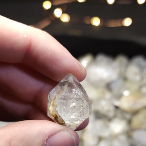 Peut inclure: Un cristal octa&eacute;drique transparent et facett&eacute; avec une inclusion dor&eacute;e. Le cristal est tenu dans une main, avec d'autres cristaux en arri&egrave;re-plan.