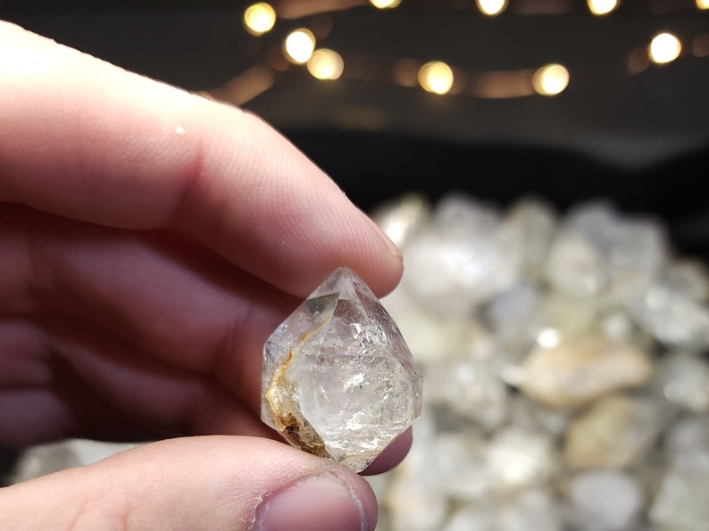 Peut inclure: Un cristal octa&eacute;drique transparent et facett&eacute; avec une inclusion dor&eacute;e. Le cristal est tenu dans une main, avec d'autres cristaux en arri&egrave;re-plan.