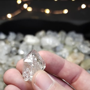 Peut inclure: Un cristal de diamant octa&eacute;drique clair et facett&eacute; avec une pointe pointue. Le cristal est tenu dans une main, avec en arri&egrave;re-plan de nombreux autres cristaux similaires.