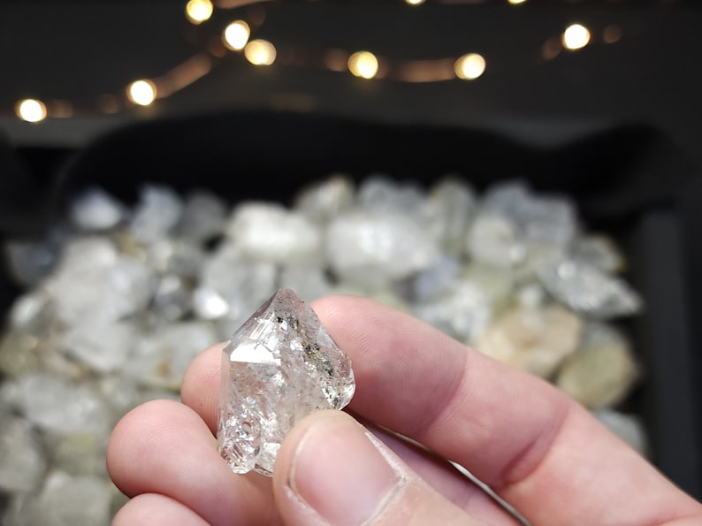 Peut inclure: Un cristal de diamant octa&eacute;drique clair et facett&eacute; avec une pointe pointue. Le cristal est tenu dans une main, avec en arri&egrave;re-plan de nombreux autres cristaux similaires.