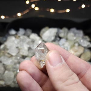 Peut inclure: Un cristal de quartz transparent avec une forme pointue, &eacute;galement connu sous le nom de diamant Herkimer. Le cristal est tenu dans une main, avec d'autres cristaux similaires en arri&egrave;re-plan.