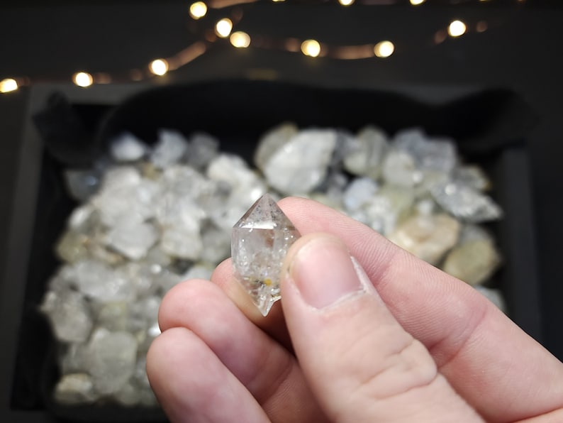 Peut inclure: Un cristal de quartz transparent avec une forme pointue, &eacute;galement connu sous le nom de diamant Herkimer. Le cristal est tenu dans une main, avec d'autres cristaux similaires en arri&egrave;re-plan.