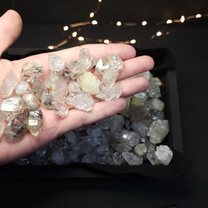 Peut inclure: Gros plan sur une main tenant une collection de cristaux de quartz transparents, facet&eacute;s et translucides. Les cristaux sont de tailles et de formes vari&eacute;es, certains ayant une l&eacute;g&egrave;re teinte jaune. Les cristaux sont dans un plateau noir avec une doublure en tissu noir doux.