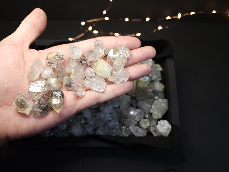 Peut inclure: Gros plan sur une main tenant une collection de cristaux de quartz transparents, facet&eacute;s et translucides. Les cristaux sont de tailles et de formes vari&eacute;es, certains ayant une l&eacute;g&egrave;re teinte jaune. Les cristaux sont dans un plateau noir avec une doublure en tissu noir doux.