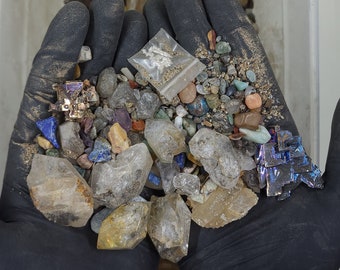 May include: A collection of various gemstones and minerals, including quartz crystals, colorful rocks, and iridescent bismuth crystals. The assortment is held in gloved hands, showcasing the natural colors and textures of the stones.
