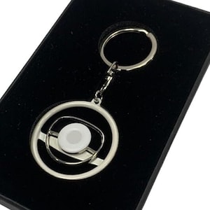 May include: A silver keychain with a spinning steering wheel design. The keychain is in a black velvet box.