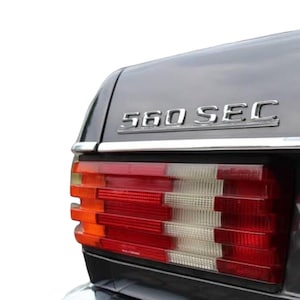 Op de afbeelding: Een close-up van de achterkant van een grijze Mercedes-Benz 560 SEC. De auto heeft een chromen sierstrip die langs de kofferbakklep loopt en een chromen bumper. De achterlichten zijn rood en wit.