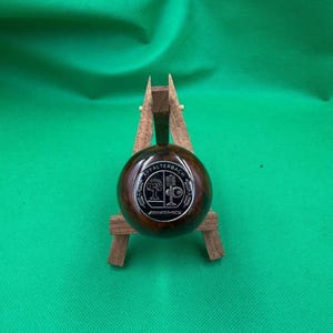 Puede incluir: Un pomo de cambio redondo, marrón oscuro, con un emblema plateado con el texto "Affalterbach" y el logotipo de AMG. El pomo se muestra sobre un pequeño soporte de madera sobre un fondo verde.