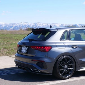May include: A grey Audi A3 hatchback with a black rear spoiler and black aftermarket wheels. The car is parked on a paved surface with a grassy field and snow-capped mountains in the background.