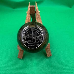 May include: A dark green, round object with a silver emblem featuring a tree and the text "AFFALTERBACH" and "AMG". The object is displayed on a small wooden stand against a green background.