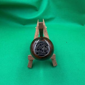 May include: A dark brown, spherical object with a silver emblem featuring a tree and the text "AFFALTERBACH" and "AMG". The object is displayed on a small wooden stand against a green background.