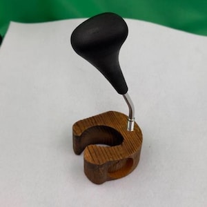 May include: A wooden espresso tamper holder with a black handle and a stainless steel base. The holder is made of a light brown wood and has a curved design to hold the tamper securely. The tamper handle is black.